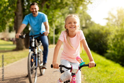 Day in the park. Happy family of caucasian father and girl riding bicycles outdoors in the summer, smiling at camera