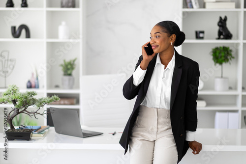 Business communication concept. Happy black businesswoman talking on cellphone standing near her workplace in office