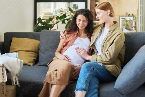 Young smiling woman touching belly of her pregnant girlfriend and embracing her while both sitting on soft comfortable couch at home
