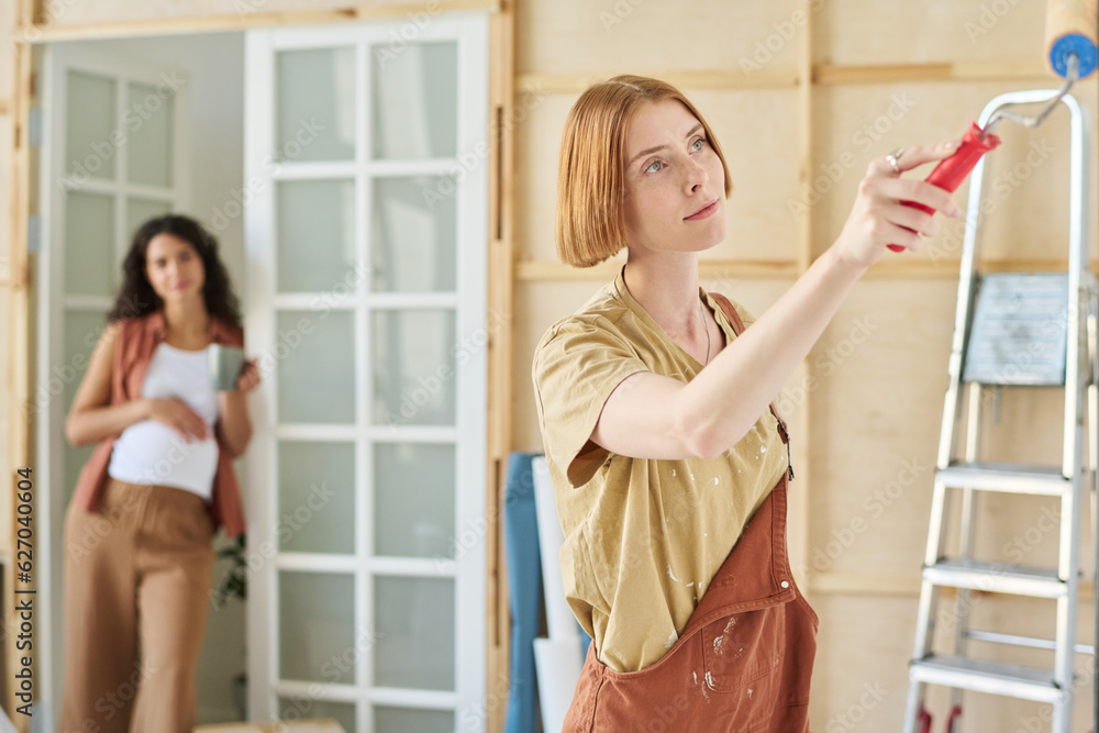 Φωτογραφία Young woman in t-shirt and coveralls applyng paint on wall of living room while