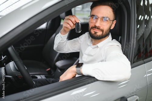 Handsome young businessman in classic white suit is smiling, looking at camera and showing car keys while sitting in car in a motor show. © Serhii