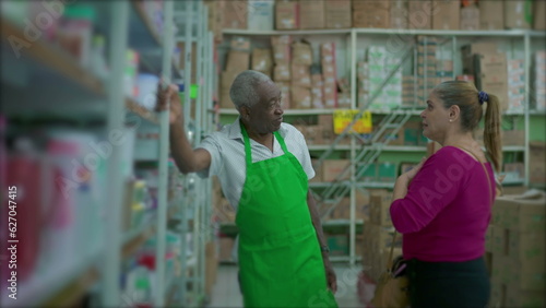 A male black senior employee listening to female customer leaning on shelf and wearing green apron, supermarket grocery store small business setting