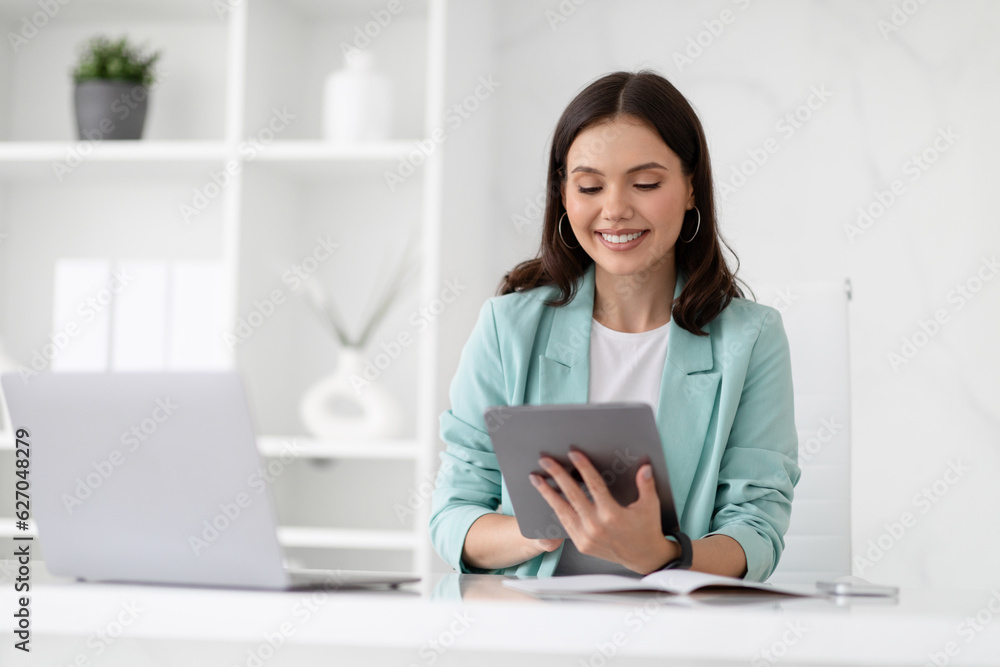 Cheerful millennial caucasian lady manager in suit typing on tablet at workplace with laptop, enjoy business Cheerful millennial caucasian lady manager in suit typing on tablet at workplace with laptop, enjoy business