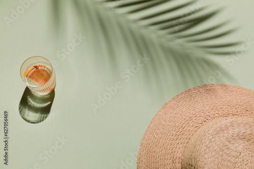 Summer flat lay with straw hat and glass of water with grapefruit slices on green background with palm leaf shadow, sun and sunlight. Vacation, holiday, summer creative minimal concept.