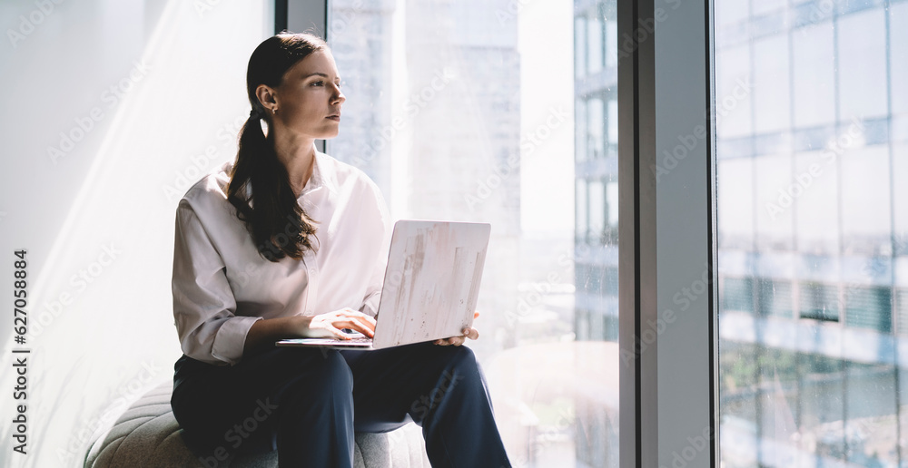 Thoughtful young woman sitting with laptop near glass wall Thoughtful young woman sitting with laptop near glass wall