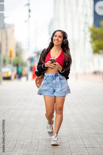 Full length young woman walking with with mobile phone and headphones on street © mimagephotos