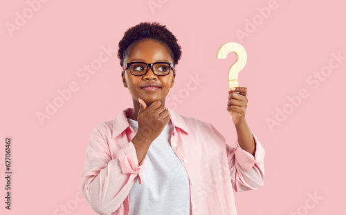 Young afroamerican woman with doubtful face in glasses and pink shirt is looking at wooden question mark in her hand on pink background. She touching chin making decision or thinking about problem.