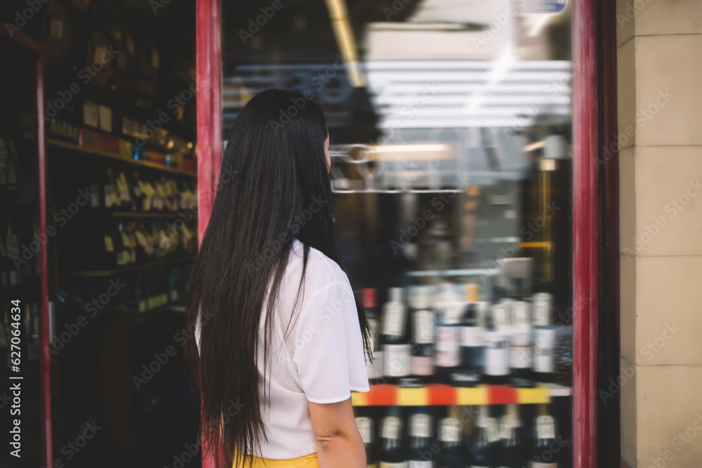 Back view of Caucasian brunette female tourist standing near shopping window and looking at showcase during travel vacations, unrecognizable woman with long dark hair visiting city downtown
