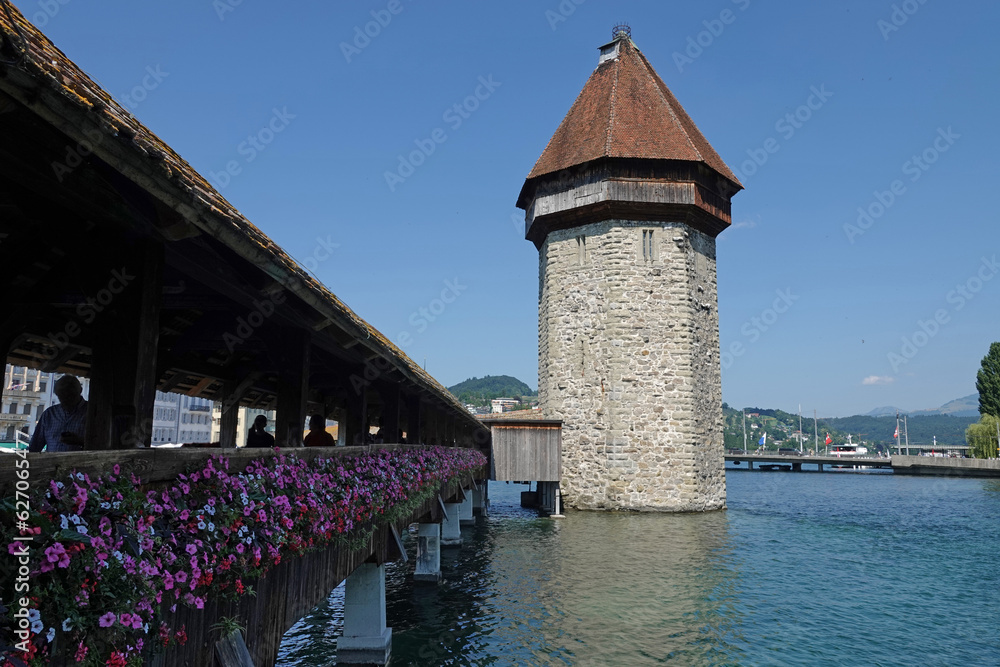 In Lucerne, Switzerland, the historic Kapellbrucke (or Chapel Bridge) and adjoining water tower are shown during the day. This bridge dates back to the 1300s and spans the Reuss River.