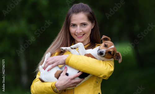 Young woman in yellow jacket holding her Jack Russell terrier dog on hands, blurred green trees background