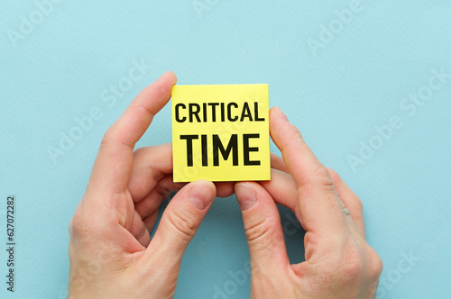 Written text CRITICAL TIME on yellow paper in hand on blue Background.