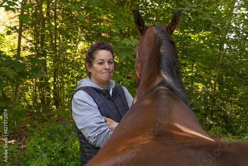 50-jährige Pferdezüchterin mit schwarzen Haaren steht nachdenklich neben ihrer hochtragenden braunen Stute im Wald und schaut in die Kamera