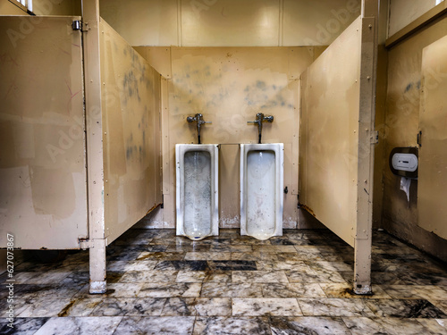 semi abandoned men's public toilet, very dirty and ruined, yellow light, dirty marble floor with various colors. Two urinals hanging on the wall, two other toilets on the sides