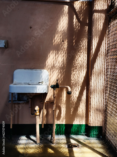 old white sink hanging on a prison wall, pipes showing, warm sunlight filtering through the security grates, creating different shapes with the shadows photo