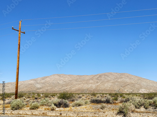 transport of electricity via high voltage cables on a wooden pole, in the middle of an empty road with earthy hills in the background, and cloudless blue sky