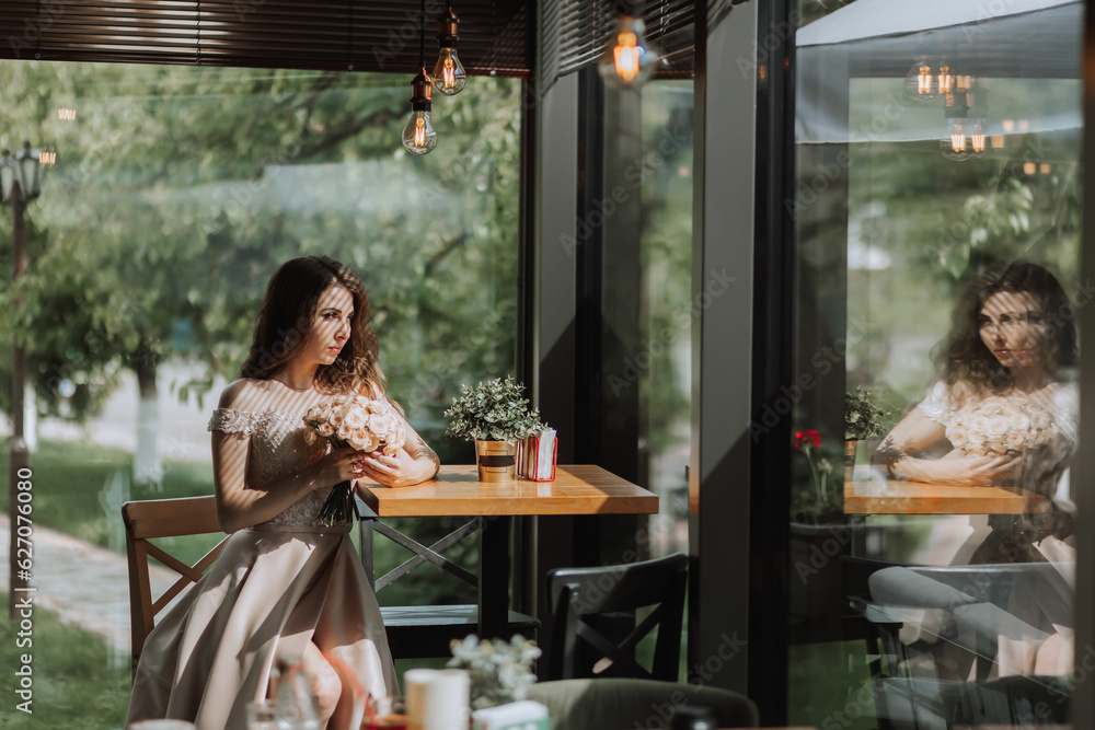 A young stylish woman in a beige dress with a bouquet of flowers in her hands is sitting in a beautiful light in a cafe in Spain A young stylish woman in a beige dress with a bouquet of flowers in her hands is sitting in a beautiful light in a cafe in Spain