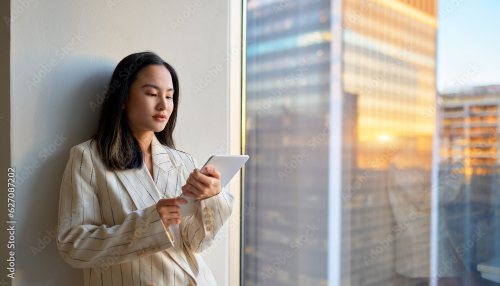 Young Asian business woman professional manager, company employee or executive using digital tablet fintech online smart tech in corporate office standing at big window, city view, copy space.