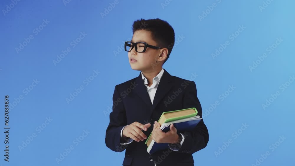 Young boy in glasses and a black suit standing with textbooks on a blue background and showing a silence gesture with his finger. Schoolboy funny pretending to be an important person