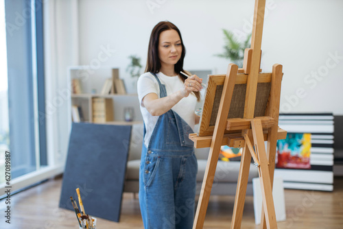 Selective focus of beautiful lady in oversized jumpsuit holding brush and palette while staying in studio. Charming young woman defining new elements on canvas with oil paints using direct lighting.