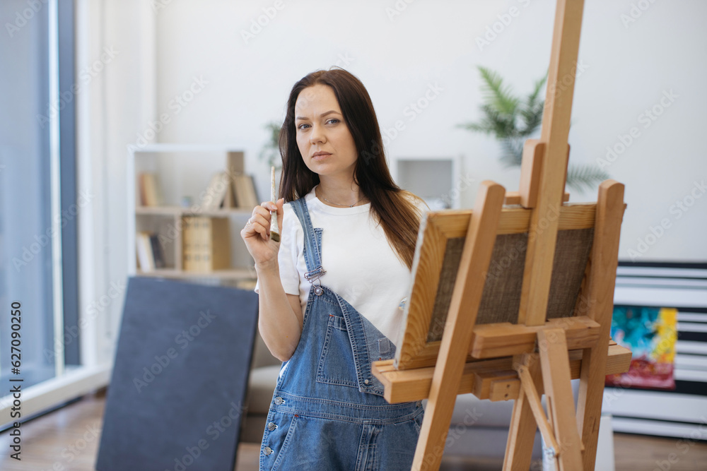 Thoughtful art person staring into distance with brush near neck while standing near wooden easel. Female professioanl in cozy overalls developing idea of future masterpiece in bright workshop. Thoughtful art person staring into distance with brush near neck while standing near wooden easel. Female professioanl in cozy overalls developing idea of future masterpiece in bright workshop.