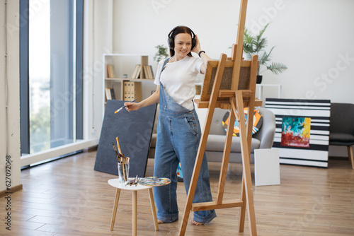 Happy brunette artist in denim jumpsuit handling brush professionally while rocking out to tunes in hands-free device. Easygoing woman approaching visual design under influence of melodies indoors.