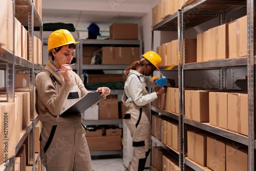 Warehouse operative looking at checklist on clipboard and counting goods boxes. Shipping company worker doing restock and checking inventory audit in distribution department stockroom photo