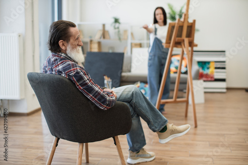 Focus on senior man in stylish wear sitting in soft chair while young woman working with canvas on easel. Relaxed model assisting inspired female by posing during life drawing lesson in workshop.
