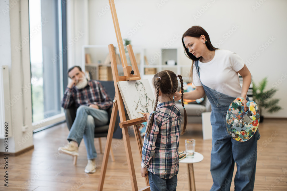Attractive woman introducing little girl to fine arts while showing sketch of senior man on canvas at home. Caring young mother helping cute daughter with color choice for grandpa's portrait.