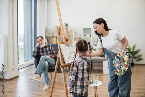 Attractive woman introducing little girl to fine arts while showing sketch of senior man on canvas at home. Caring young mother helping cute daughter with color choice for grandpa's portrait.