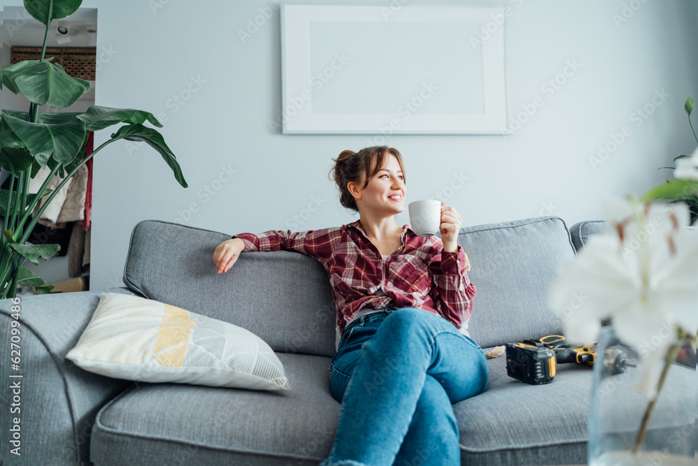 Happy smiling proud of herself young woman relaxing with coffee cup after she puts by her own a picture on the wall at her home. Housekeeping work. Doing repair herself. DIY, gender equality concept. Happy smiling proud of herself young woman relaxing with coffee cup after she puts by her own a picture on the wall at her home. Housekeeping work. Doing repair herself. DIY, gender equality concept.