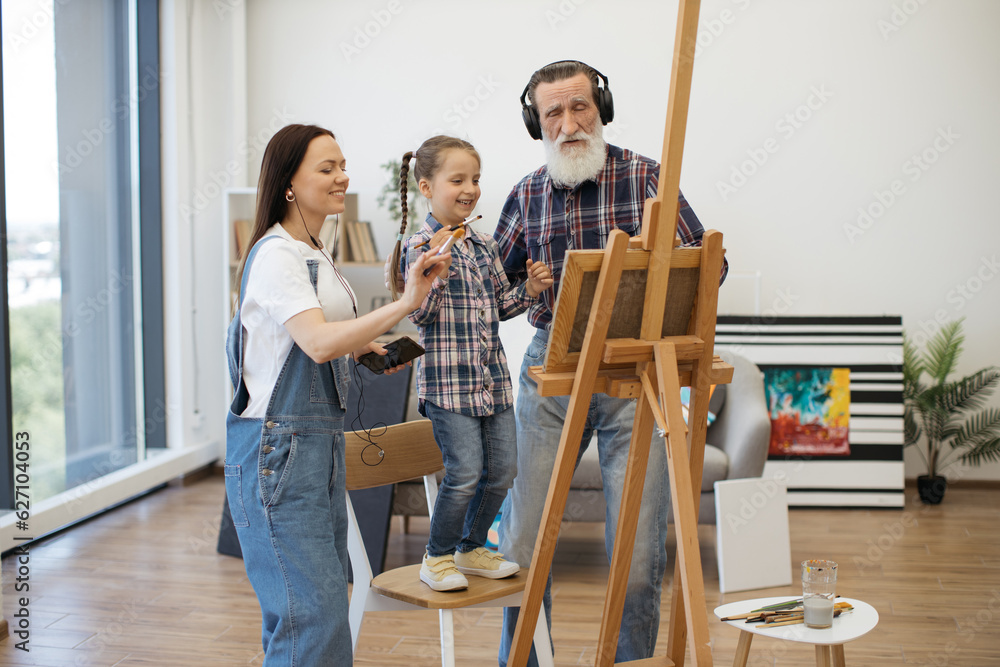 Modern pensioner in headphones and smiling lady with wired earbuds dancing with cute girl while working on canvas. Artistic family finding inspiration in music while creating picture in living room. Modern pensioner in headphones and smiling lady with wired earbuds dancing with cute girl while working on canvas. Artistic family finding inspiration in music while creating picture in living room.