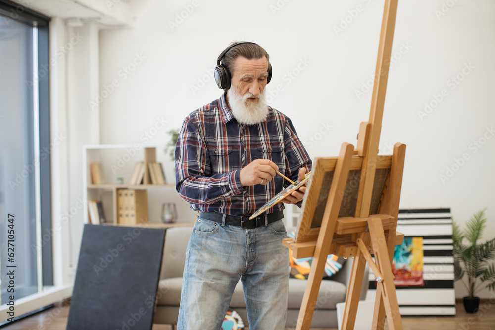 Portrait of smiling pensioner creating artwork while listening to songs via wireless device in room interior. Passionate painter in cozy clothes living full life of inspiration in retirement.