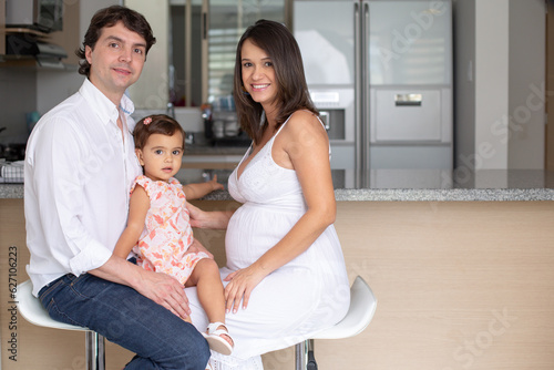 Smiling married pregnant couple at home, carrying their daughter, sitting on benches at the kitchen counter inside their home.