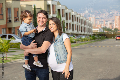 Happy smiling pregnant family standing in a street of a residential area with father holding their daughter in arms and hugging.