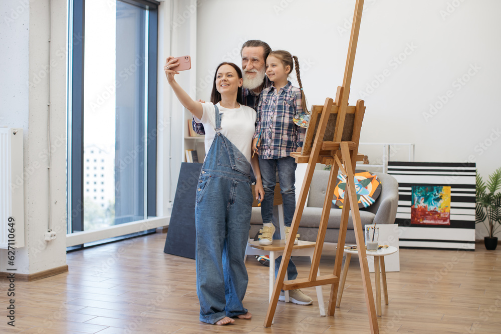 Happy family of three painters making selfie on modern cellphone while working on new drawing together at home studio. Loving people enjoying spending pastime with each other.