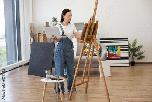 Female professional painter having video conference using portable computer in comfy flat with amazing panoramic view. Attractive female in casual outfit having online art class with beginners.