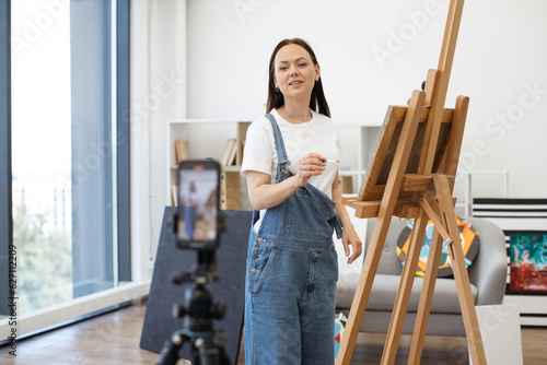 Talented female artist using tripod with cellphone for making online broadcast for own subscribers while holding art courses. Charming long-haired brunette standing near easel in spacious living room.