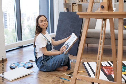Side view of talented artist sitting on floor in lotus position with canvas on knees and drawing with brush. Beautiful lady with long brown hair painting portrait of man at comfortable home studio.