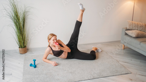 Calorie burn training. Weight loss fitness workout. Relaxed athletic woman eating sweet doughnut dessert doing physical exercise on floor at home interior with free space.