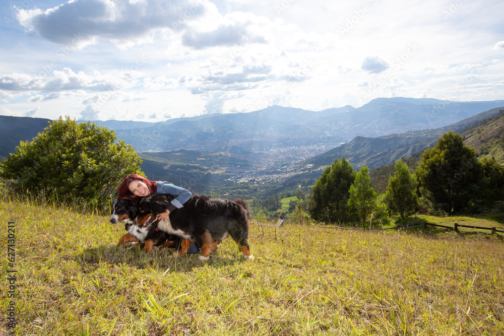 Young beautiful redhead woman hugging her two big Bernese Mountain dogs in a mountain landscape with mountains in the background and a city in the valley below.