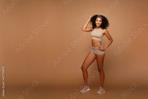 A beautiful ebony model in a sporty slim top and shorts stands on a beige background with copy space.