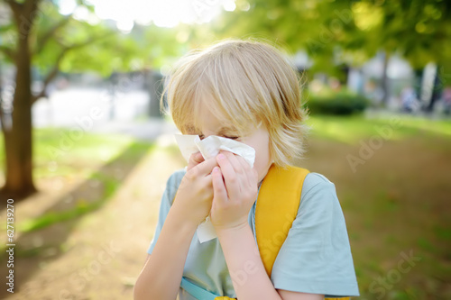 Preteen boy sneezing and wipes nose with napkin during walking in summer park. Flu season and cold rhinitis. Allergic kid.