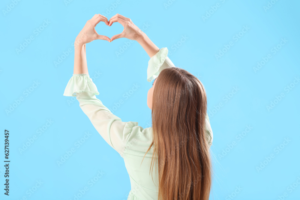 Young woman in dress making heart shape with hands on blue background, back view Young woman in dress making heart shape with hands on blue background, back view