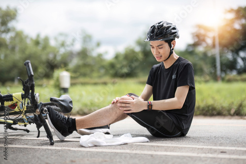 An injured young Asian male cyclist in sportswear and a bike helmet fell off the bike while biking along country roads. knee pain, knee bleeding, injured knee