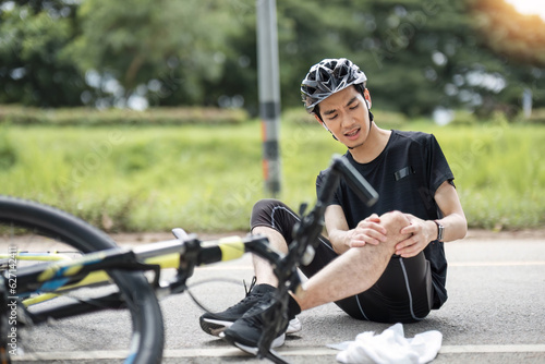 An injured young Asian male cyclist in sportswear and a bike helmet fell off the bike while biking along country roads. knee pain, knee bleeding, injured knee