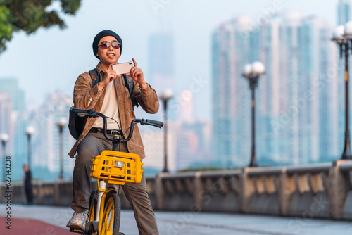 Asian man is riding a bike and using mobile phone to take photo outdoor in the city. 