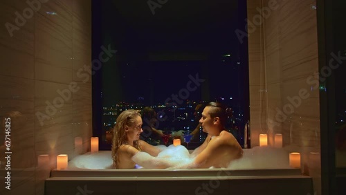 Love couple enjoys romantic honeymoon in hotel. Young married couple relax in bathtub with view of city night. Embracing in foam, kiss and hug.Honeymoon vacation in romantic atmosphere in candlelight photo