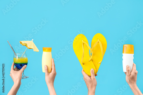 Female hands with bottles of sunscreen cream, cocktail and flip flops on blue background