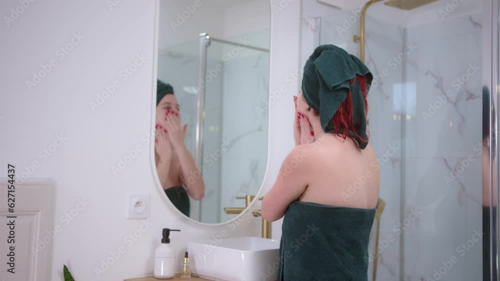 Relaxed red hair girl skin caring the face in front of the mirror in a modern bathroom. She is wearing a green turban. After shower care. Half-lenght shot and slow mo Relaxed red hair girl skin caring the face in front of the mirror in a modern bathroom. She is wearing a green turban. After shower care. Half-lenght shot and slow mo