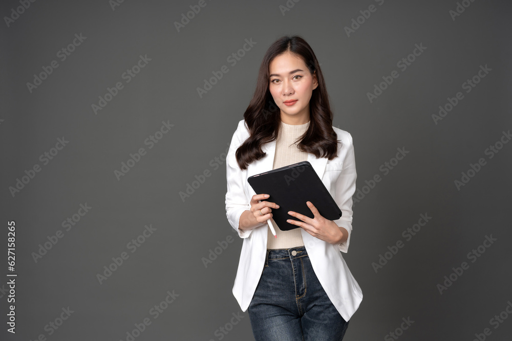 Long-haired Asian female executive posing in a white suit, jeans, holding a tablet in her hand and a pen, making eye contact, and taking pictures in a gray background studio. Long-haired Asian female executive posing in a white suit, jeans, holding a tablet in her hand and a pen, making eye contact, and taking pictures in a gray background studio.
